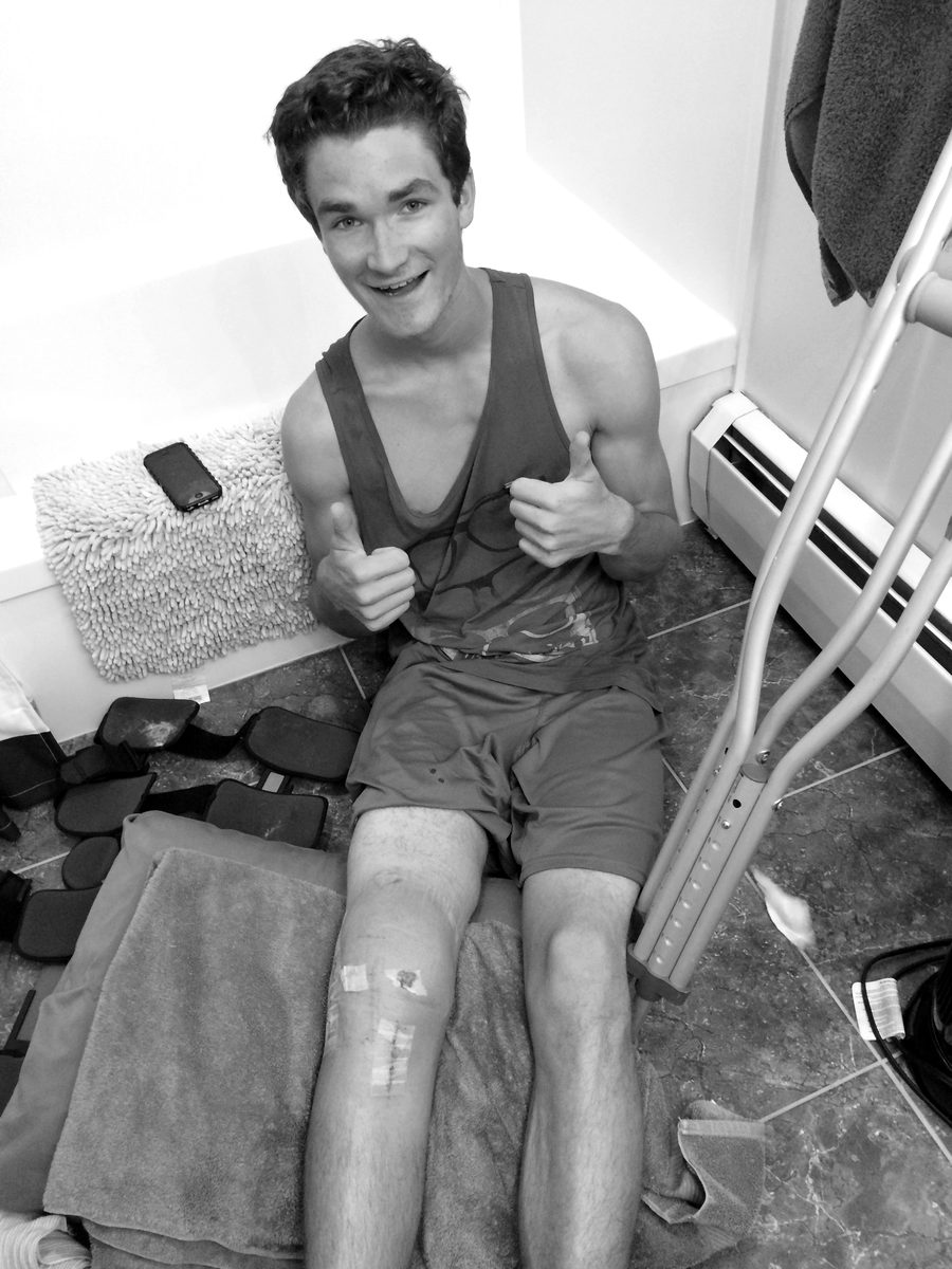 Connor on the bathroom floor two days after his second reconstruction, bandaged right knee, crutches beside him, two thumbs up