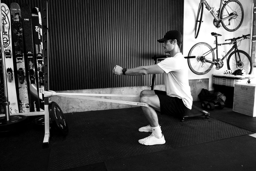 Connor doing a band-resisted squat holding a plate in his garage gym, skis and bikes on the wall behind him