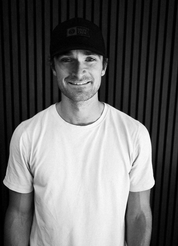 Connor Clayton in a black cap and white tee against the slat wall in his garage gym, smiling at the camera