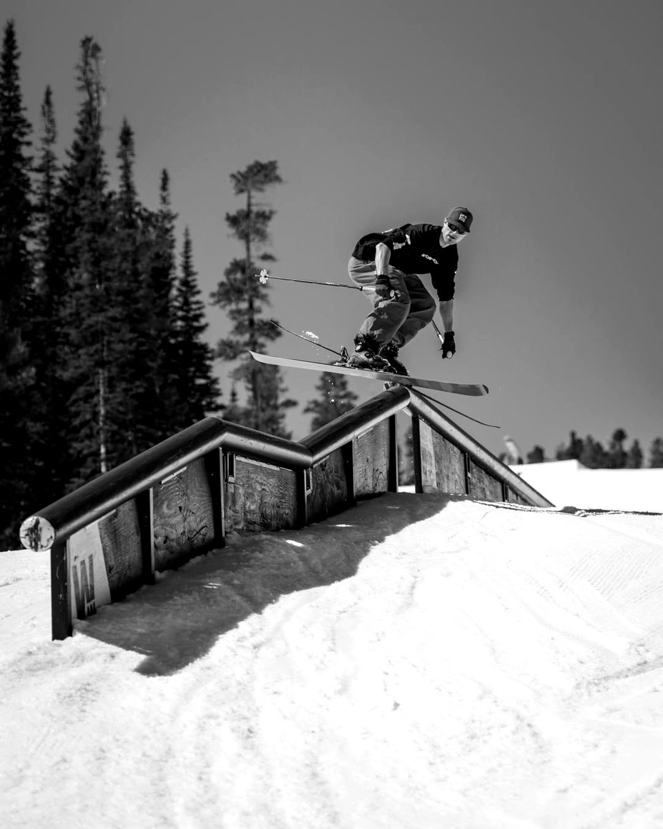 Connor sliding a long down rail in a terrain park, skis crossed over the box, blue sky and pines behind