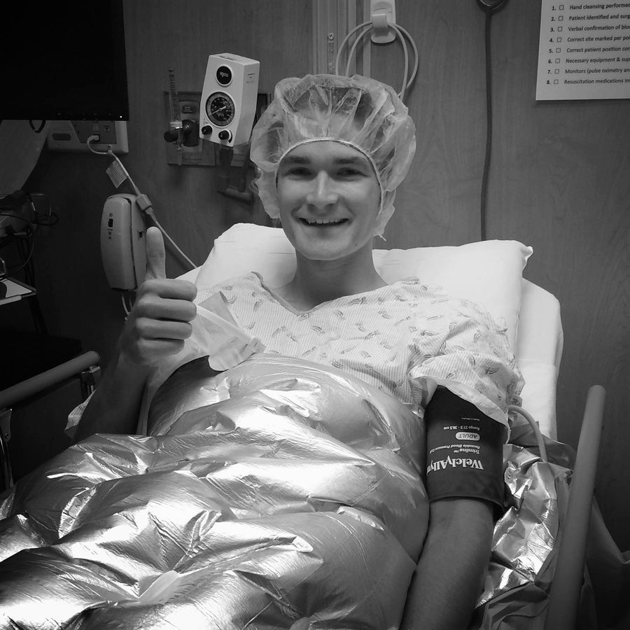 Connor in a hospital bed in a surgical cap and gown, thumbs up before ACL surgery