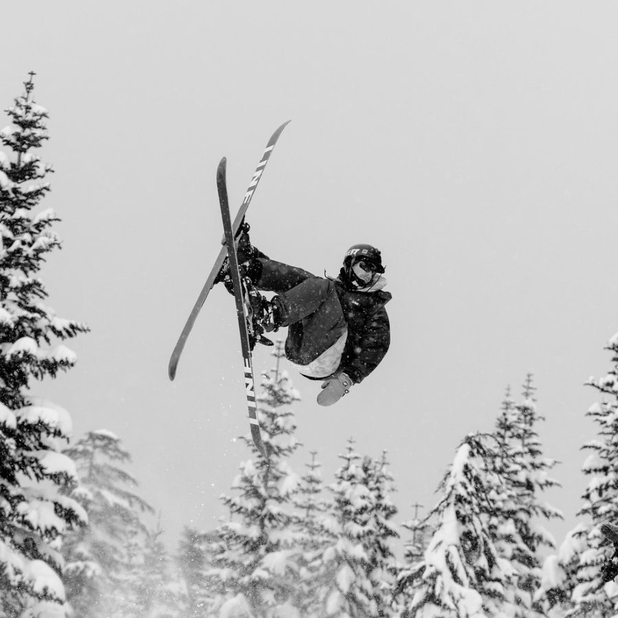 Connor upside down mid flip on skis above a snowy jump, a filmer below